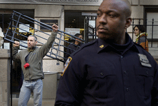 Tim Keating, wearing a green fleece jacket, with a heavy bicycle chain around his neck, locked to a black steel gate door that is wedged horizontally across the gate opening, the Mexican Consolate in the background, with a police officer to the right.